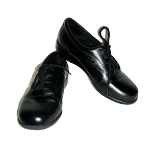 May include: A pair of black leather lace-up shoes. The shoes have a low heel and a glossy finish. The laces are black and tied in a bow. The shoes are set against a white background.