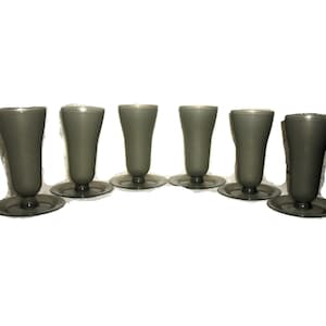 May include: Six gray-tinted glass goblets with a flared top and a circular base are arranged in a row. The stemware has a simple, elegant design, suitable for serving beverages. The glasses are evenly spaced against a white background.