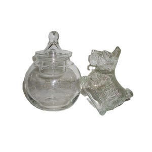 May include: A clear glass jar with a lid and a decorative glass dog figurine. The jar is round with a small handle on top. The dog is a Scottish Terrier. Both items are on a white background.