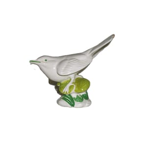May include: A white ceramic bird figurine with a green beak and eye details. The bird is perched on a base with green and yellow leaves. The figurine has a glossy finish and is likely a decorative item.
