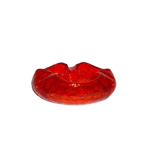 May include: A vibrant red glass ashtray featuring a textured, bubbled surface throughout the material. The circular object has a distinctively wavy, scalloped rim with multiple indentations designed for resting items along the top edge.