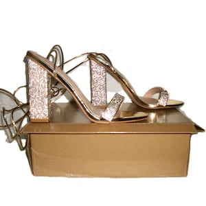 May include: Gold-colored high-heeled sandals with rhinestone embellishments on the block heels and straps. The sandals are displayed on a gold-colored box, with delicate straps extending from the heels.