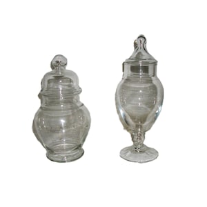 May include: Two clear glass apothecary jars with lids. The jar on the left has a rounded body and a bulbous lid. The jar on the right has a more elongated shape, a pedestal base, and a decorative lid. Both jars are empty.