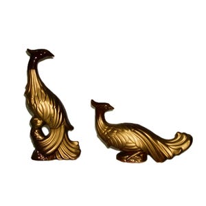 May include: Two gold ceramic bird figurines. The larger bird is standing upright with its wings spread out. The smaller bird is lying down with its wings spread out. Both birds have brown accents on their heads and tails.