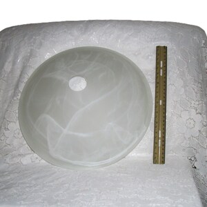 May include: A circular, frosted white glass light shade featuring a swirling, marbled pattern and a central circular opening. A wooden ruler is positioned vertically next to the shade, indicating a diameter of approximately 12 inches.