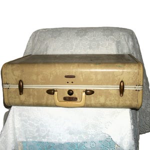 May include: Vintage beige suitcase with brass hardware, a cream-colored handle, and a white stripe. The suitcase has a rectangular shape and is resting on a white lace tablecloth. The suitcase is a classic travel accessory.