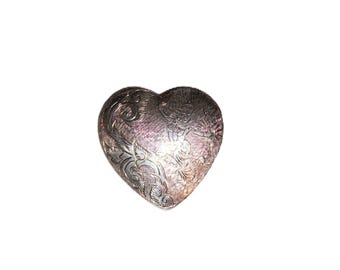 May include: A silver heart-shaped trinket box with intricate floral and figure engravings. The box has a slightly textured surface and a polished finish, reflecting light. The design features detailed patterns and a secure closure.