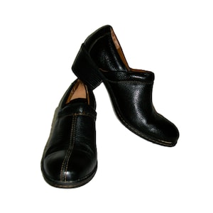 May include: A pair of black leather clogs with a low heel. The shoes have a textured surface and a stitched seam down the center of the vamp. The heels are approximately 2 inches high.