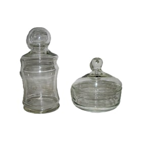 May include: Two clear glass jars with lids. One is tall and cylindrical, the other is shorter and wider. Both have rounded lids and are empty. The jars are made of transparent glass.