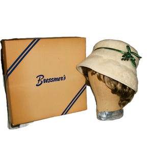 May include: A vintage cream-colored cloche hat with a green ribbon and embellishment, displayed on a mannequin head. Beside it is a tan box with the name "Bressmer's" printed on it, with blue diagonal stripes.
