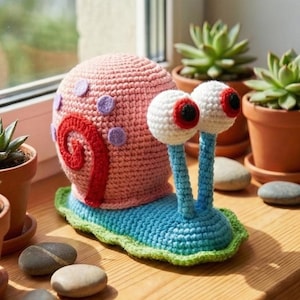 May include: A handmade crochet snail with a pink shell, red swirl, and purple spots. The snail has large white eyes with red details and blue eye stalks. It sits on a blue base with a green border. The snail is displayed on a wooden surface with potted succulents.
