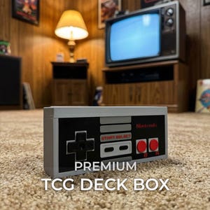 Op de afbeelding: Een rechthoekige grijze, zwarte en rode deck box ontworpen om op een vintage videogamecontroller te lijken. De voorkant heeft een zwart paneel met het Nintendo-logo, Start- en Select-knoppen, een richtingspad en twee rode A- en B-knoppen.