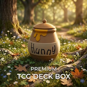 May include: A tan, round TCG deck box shaped like a honey pot with a lid, featuring a yellow honey drip design on the top rim and the word "Hunny" written in black script on the side, set against a blurred forest floor with fallen leaves.