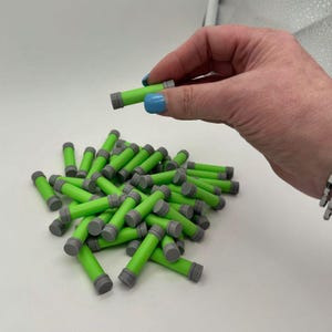 Puede incluir: Una colección de objetos cilíndricos verde neón y grises. Uno se sostiene entre los dedos, con muchos más dispersos debajo. Los objetos tienen un cuerpo verde brillante y extremos grises. El fondo es una superficie blanca lisa.