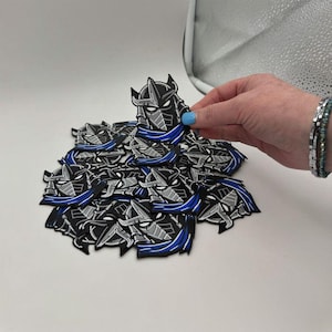 May include: A pile of black, gray, and blue patches featuring a character's helmet design. The patches are on a white surface. A hand with blue nail polish holds one patch.