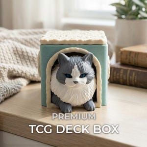 May include: A square, light blue deck box featuring a sculpted gray and white cat with blue eyes peeking out from an arched opening. The top of the box has a textured, cream-colored surface. The text "PREMIUM TCG DECK BOX" is printed below.