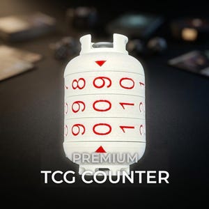 May include: A white cylindrical TCG counter featuring three rotating dials with red numbers and red triangular indicators. The device displays the text PREMIUM TCG COUNTER in white capital letters at the bottom, designed for tracking game scores.