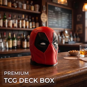 May include: A red and black TCG deck box shaped like a superhero mask with white eye accents and a textured surface. The deck box is positioned on a wooden surface. The text PREMIUM TCG DECK BOX is visible in the lower left corner of the image.