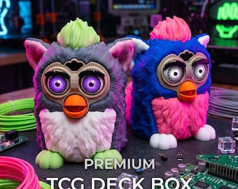 Puede incluir: Dos cajas para mazos TCG con forma de criatura, pelaje texturizado, orejas puntiagudas y picos naranjas. La caja izquierda es gris con un mechón verde y ojos morados, la derecha es azul con un mechón rosa y ojos blancos. Texto: PREMIUM TCG DECK BOX.