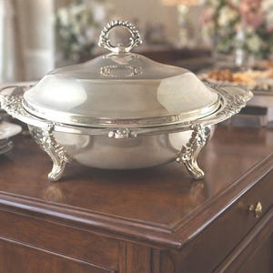 Puede incluir: Una fuente de plata con tapa y detalles ornamentados, sobre un aparador de madera oscura. El plato tiene asas decorativas y se apoya en tres patas curvas. Otros platos y vajilla se ven al fondo.