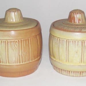 May include: Two ceramic barrel-shaped containers with lids and handles. The containers are tan and brown with vertical wood grain detailing. The lids have a knob on top. The handles are curved.