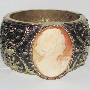 May include: A gold-toned hinged bangle bracelet with a black enamel design and a central cameo. The cameo features a profile of a woman in cream and peach tones. The bracelet has a detailed, ornate pattern.