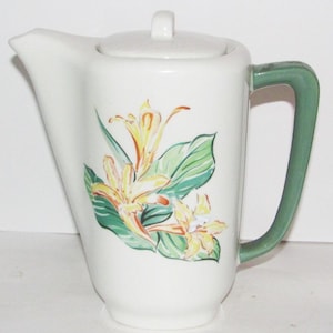 May include: White ceramic coffee pot with a green handle. The pot features a floral design with yellow and orange flowers and green leaves. The lid has a small white knob.