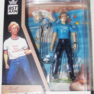 May include: A Napoleon Dynamite action figure in a clear plastic package. The figure wears a blue shirt and dark pants. The packaging includes the text "Napoleon Dynamite" and "Vote for Pedro."