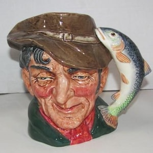 May include: A ceramic mug shaped like a man's head, wearing a brown hat. A fish is attached to the side of the mug. The mug has a green collar and a red shirt. The man's face is detailed with wrinkles and rosy cheeks.