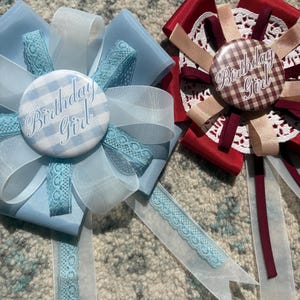 May include: Two decorative birthday badges. One is light blue with a gingham center and the words "Birthday Girl". The other is red with a gingham center and the words "Birthday Girl". Both have ribbons and lace.