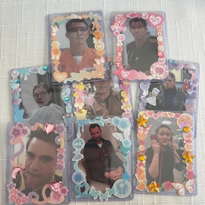 May include: Seven rectangular plastic card holders containing photographs, each decorated with colourful stickers, gems, and charms. The frames feature various motifs including hearts, stars, butterflies, and bubbles in pastel shades, surrounding images of different individuals.