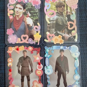 May include: Four rectangular trading cards in clear plastic sleeves, each featuring a character from the television series Merlin. The cards are decorated with colorful stickers including hearts, stars, flowers, and pearls in various pastel shades.