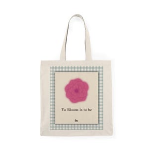 Puede incluir: Un bolso de tela beige con un gráfico central rectangular con un borde de cuadros verdes y blancos. El gráfico muestra una ilustración de una flor rosa vibrante sobre el texto negro To Bloom is to be y un pequeño logotipo en la parte inferior.