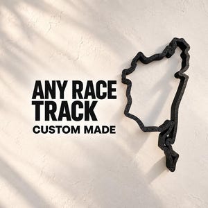 Puede incluir: Un contorno negro tridimensional de una pista de carreras montado sobre una superficie texturizada. A la izquierda de la pista, el texto negro dice: ANY RACE TRACK CUSTOM MADE. El objeto presenta un acabado mate rugoso y una forma irregular.