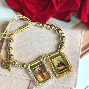 May include: A bracelet featuring alternating gold and silver-toned beads with two rectangular pendants. One pendant displays an image of Jesus, and the other shows the Virgin Mary with the infant Jesus. A small circular charm is attached to the side.