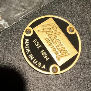 May include: A round, black and gold Gibson Custom guitar part. The gold-colored center features the Gibson logo and the text "EST. 1894 MADE IN U.S.A." Four holes are drilled around the edge. A small bag of screws is in the upper left.