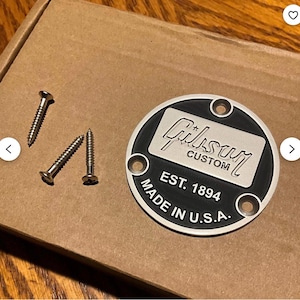 May include: A black and silver Gibson Custom plate with the text "Gibson Custom" and "EST. 1894 MADE IN U.S.A." is shown. Three silver screws are included. The plate is round with three screw holes and a silver border.