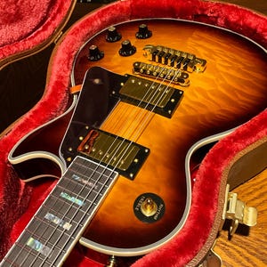 Può includere: Una chitarra elettrica con finitura sunburst e hardware dorato, in una custodia aperta foderata in velluto rosso. La chitarra presenta manopole nere, una tastiera con intarsi in madreperla e un selettore "Treble" e "Rhythm". La custodia è marrone con una chiusura dorata.