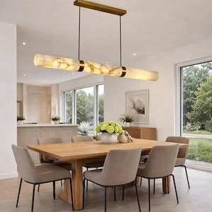May include: A modern dining room with a rectangular wooden table and six upholstered chairs. Above the table hangs a linear chandelier with three cylindrical, marble-like shades and gold accents. The room has large windows and a neutral color palette.