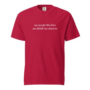 May include: A red t-shirt featuring the white text "we accept the love we think we deserve". The shirt has a crew neck and short sleeves. The garment is a solid colour and appears to be made from a soft fabric, suitable for casual wear.