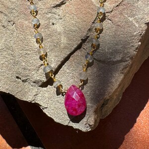 May include: A gold chain necklace with a pink gemstone pendant. The chain is made of small, round beads and gold links. The pendant is a teardrop shape and is set in a gold setting.
