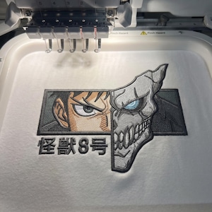 May include: An embroidery design on white fabric featuring a split-face character graphic. One side shows a human eye, the other a grey monster mask with a blue eye. Below the graphic, the black Japanese text reads Kaiju No. 8 in a bold font.