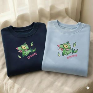 May include: Two crew neck sweatshirts, one navy blue and one light blue. Each features an embroidered green cartoon creature with a pink bow and the name "Dreepy" in pink script. The sweatshirts are made of a soft material.
