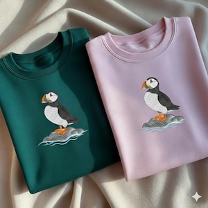 Può includere: Due felpe, una verde smeraldo e una rosa chiaro, ciascuna ricamata con un disegno di pulcinella di mare. Le pulcinelle di mare sono raffigurate su una roccia con dettagli d'acqua, con colori nero, bianco, arancione e grigio.