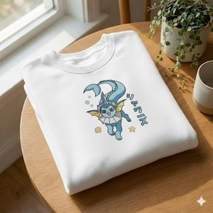 以下が含まれることがあります： 青と黄色の生き物の漫画イラストと日本語のテキストが特徴の白いクルーネックのトレーナー。デザインには泡と星が含まれています。トレーナーは木製の表面に折りたたまれています。