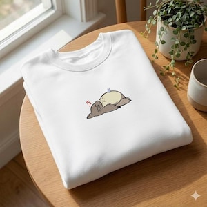 May include: White crew neck sweatshirt featuring a cartoon illustration of a sleeping creature. The illustration includes a light brown creature with a cream-colored belly and a small blue butterfly. The word "N" with a few z's is next to the creature.