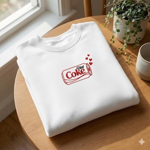 Può includere: Felpa bianca con un design ricamato rosso di una lattina di Diet Coke e piccoli cuori rossi. La felpa è realizzata in un materiale morbido ed è esposta su un tavolo di legno. Il design è una rivisitazione giocosa di una bibita classica.