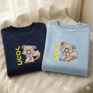 May include: Two sweatshirts, one navy blue and one light blue, each featuring a cartoon dog graphic and Japanese text in yellow. The dog has brown and tan fur with a white chest and a smiling expression. The sweatshirts are displayed on a neutral background.