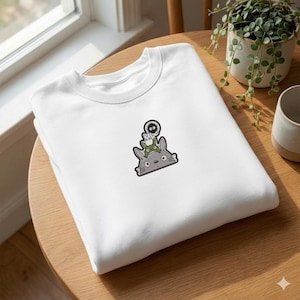 May include: A white crewneck sweatshirt featuring a small, centred embroidered graphic of a large grey creature with pointed ears, topped by a smaller green creature, a tiny white creature, and a small black fuzzy soot sprite character.
