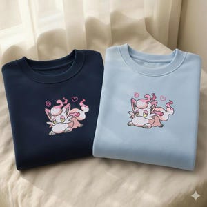 Puede incluir: Dos sudaderas de cuello redondo, una azul marino y otra azul claro. Cada sudadera presenta un zorro de dibujos animados bordado con detalles en rosa y blanco, incluyendo remolinos y corazones. Las sudaderas se muestran sobre una superficie color crema.